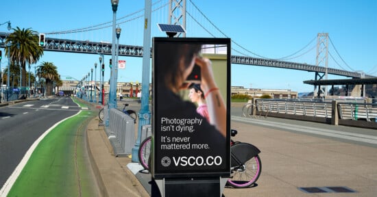 A street advertisement near a bridge promotes VSCO with a photo of a person holding a camera. The sign reads, "Photography isn't dying. It's never mattered more. VSCO.CO." A bike lane and city background are visible.
