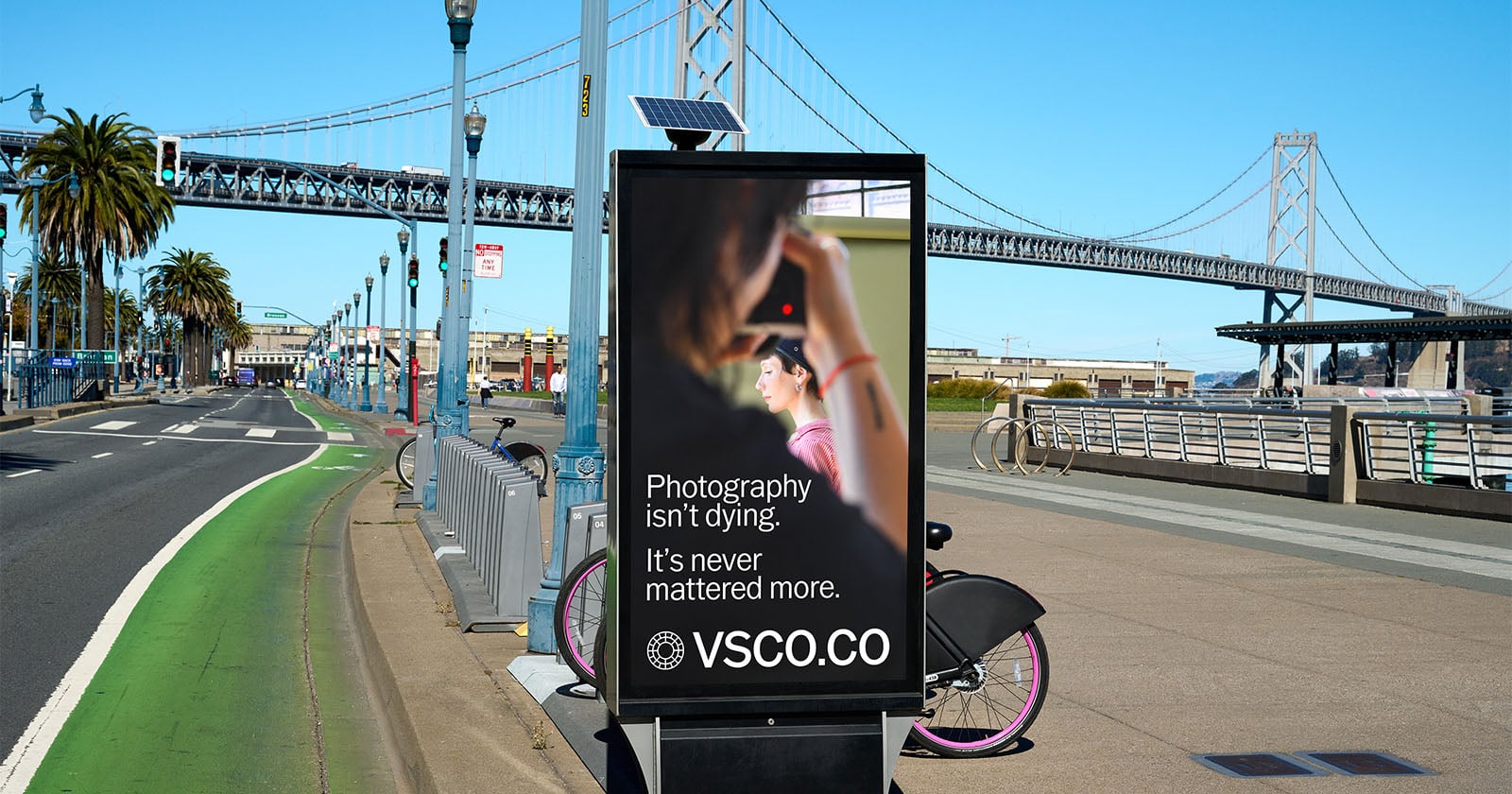 A street advertisement near a bridge promotes VSCO with a photo of a person holding a camera. The sign reads, "Photography isn't dying. It's never mattered more. VSCO.CO." A bike lane and city background are visible.