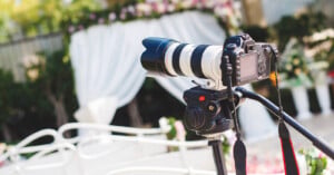 A professional camera with a large zoom lens is mounted on a tripod, set up outdoors in front of a white wedding arch decorated with flowers and draped fabric.