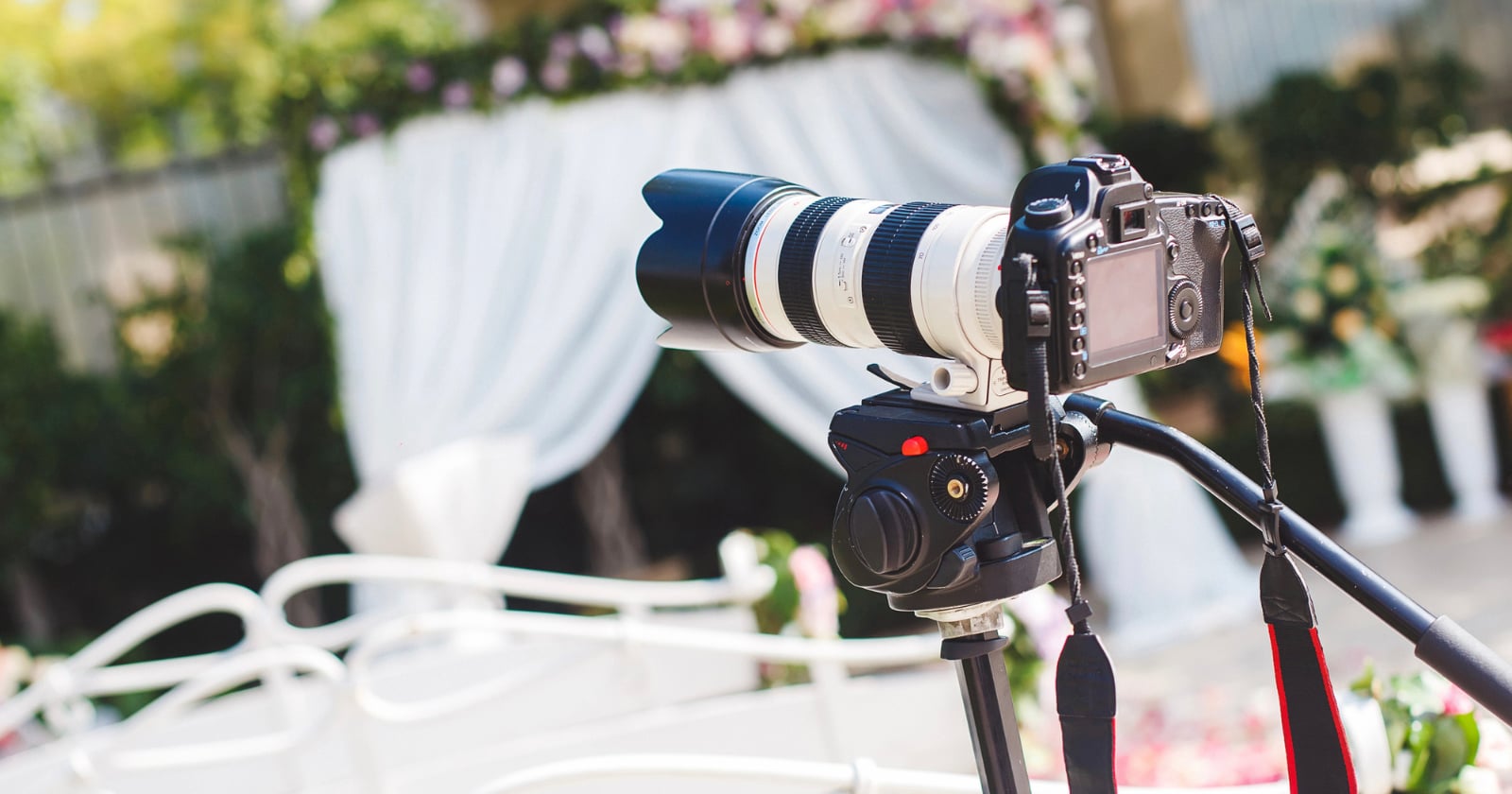 A professional camera with a large zoom lens is mounted on a tripod, set up outdoors in front of a white wedding arch decorated with flowers and draped fabric.