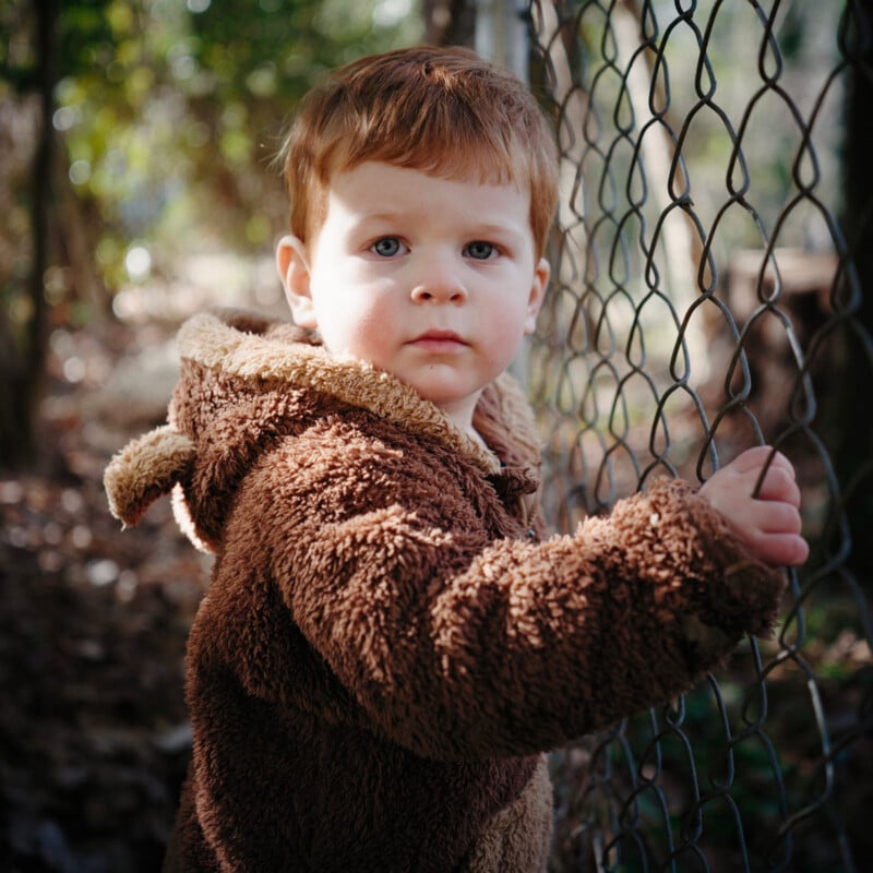 A young child with short brown hair wearing a fuzzy brown jacket stands by a chain-link fence, looking at the camera. Sunlight filters through trees in the background, creating a natural outdoor setting.