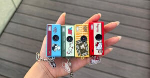 A hand with long, manicured nails holds four small toy cameras with different colorful designs, including Hello Kitty and playful patterns, each with a matching keychain attached. The background is wooden planks.