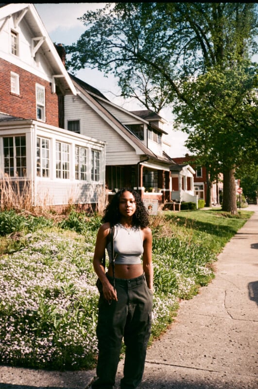 A young woman with curly hair, wearing a gray crop top and dark pants, stands on a sunny sidewalk in front of houses with green lawns and trees.