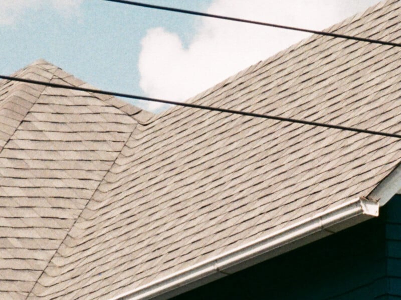 Close-up of a gray shingled roof with white gutters against a blue sky with clouds. Three black power lines run diagonally across the upper part of the image.