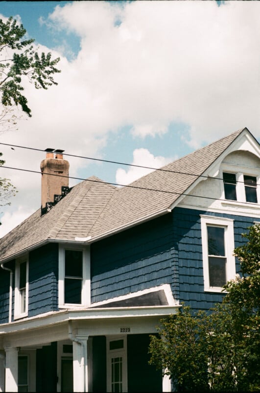 A two-story house with blue siding, white trim, and a gray shingled roof is pictured under a partly cloudy sky. A brick chimney and large windows are visible, with trees framing the home.
