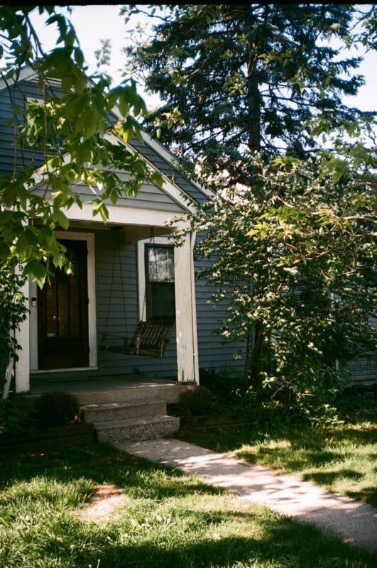 A small gray house with a covered porch, a wooden swing hanging by the door, green grass, and leafy trees casting shadows in the sunlight.