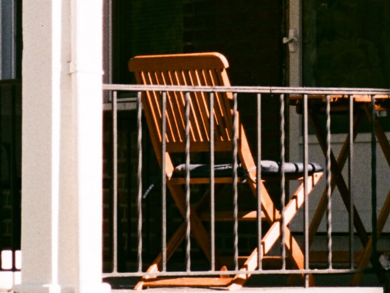 A wooden chair with a dark cushion sits behind a metal railing on a sunlit balcony or porch, next to a matching wooden table.