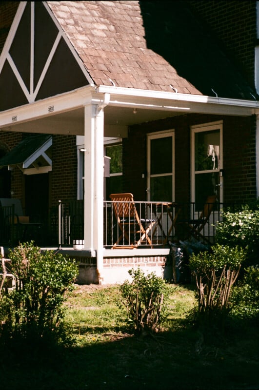 A wooden rocking chair sits on a small porch of a brick house, partially shaded by the roof. Green bushes line the front yard under bright sunlight.