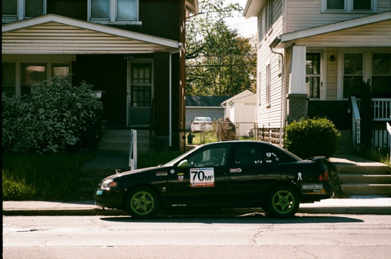 A black sports car with green rims and racing decals, including "70MF," is parked on the street in front of two houses with porches and stairs, on a sunny day.