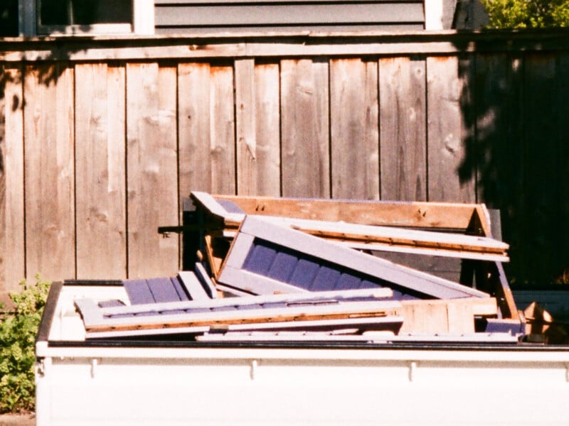 A pile of dismantled blue and wooden panels lies in the back of a white pickup truck parked in front of a weathered wooden fence.
