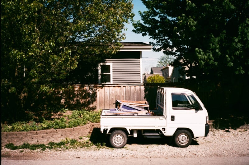 A small white pickup truck parked on a sunlit street in front of a wooden fence and a house, with various items loaded in its bed. Lush green trees and bushes surround the area.