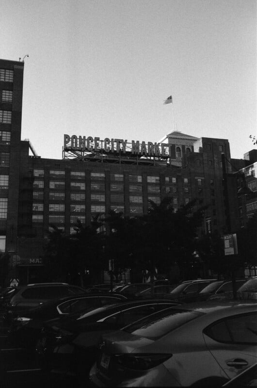 A black-and-white photo shows cars parked in front of the Ponce City Market building, with its large rooftop sign and an American flag visible against a clear sky.