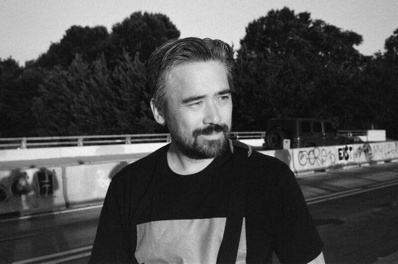 Black and white photo of a man with short hair and a beard standing outdoors near a graffiti-covered wall, with trees in the background and sunlight casting shadows on his face.