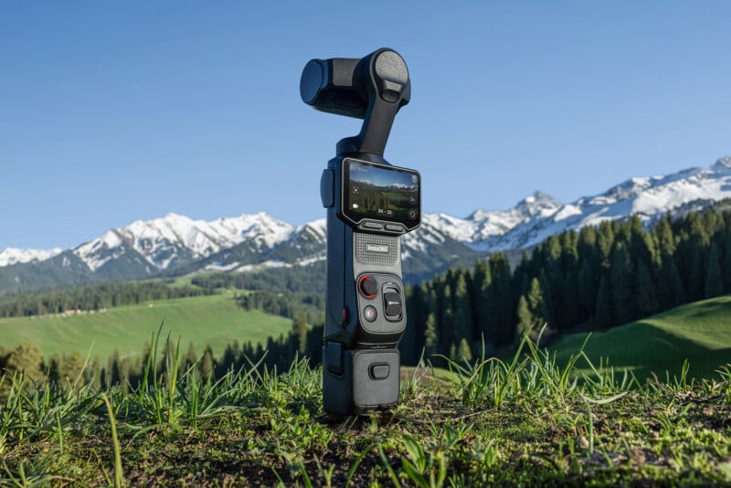 A handheld camera gimbal with a mounted screen stands on grass, with snow-capped mountains, green hills, and a blue sky in the background.