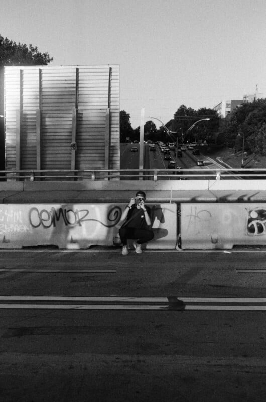 A person sits alone on a graffiti-covered overpass wall, holding a camera and facing the street. Cars drive below and trees line the road in the background. The image is in black and white.