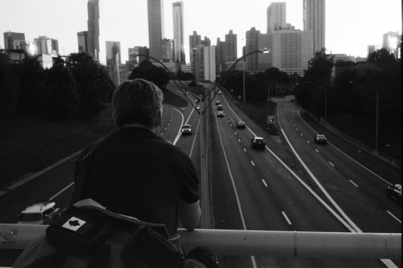 A person with a backpack featuring a Canadian flag patch leans on a bridge railing, overlooking a multi-lane highway with cars and a city skyline in the distance at dusk.