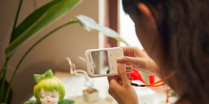 A person holds a small white digital camera, pointing it at a green-haired doll surrounded by potted plants on a table near a window.