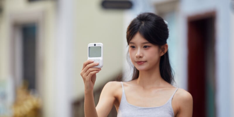 A young woman with long dark hair holds up a small white electronic device, possibly a camera or medical monitor, while standing outdoors and looking at it.