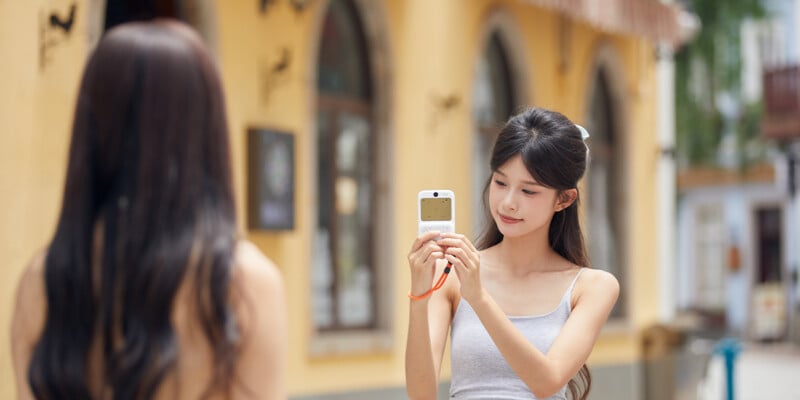 A young woman outdoors holds a small camera, taking a photo of another woman with long hair standing in front of her. The background features arched windows and a yellow building.