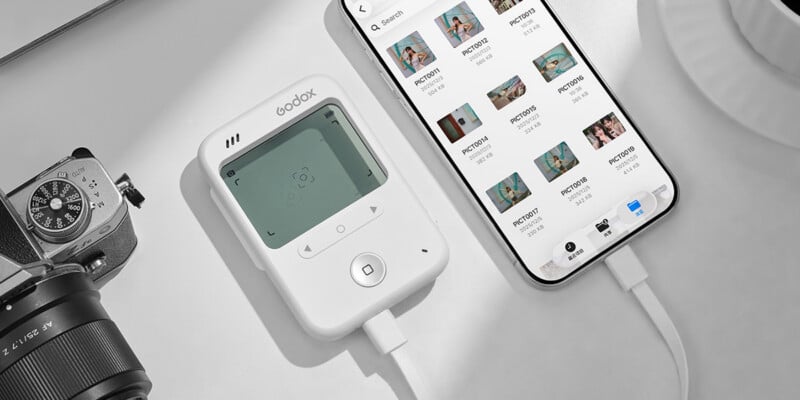 A Godox device and a smartphone connected by a cable rest on a white surface near a vintage camera and a cup; the phone screen displays a photo gallery app with image thumbnails.
