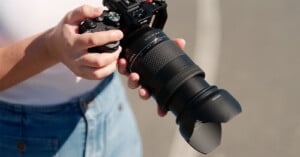 A person holding a professional DSLR camera with a large zoom lens, wearing a white shirt and blue jeans, standing outdoors on a paved surface.