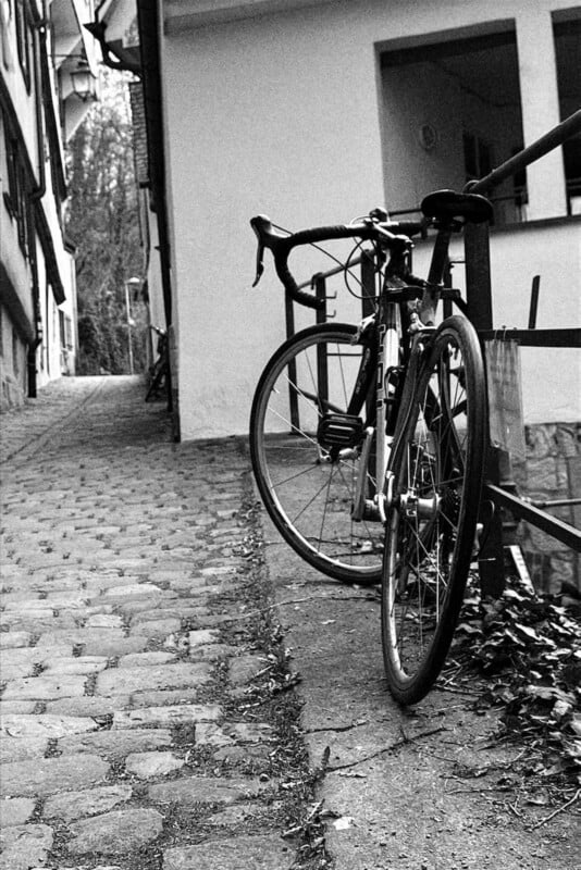 A road bicycle is locked to a railing beside a cobblestone street lined with buildings. The image is in black and white, giving it a vintage, nostalgic feel.