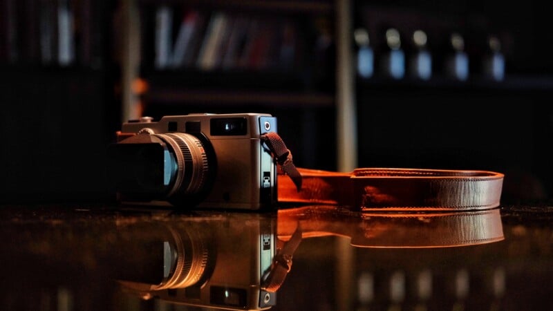 A vintage camera with a brown leather strap rests on a glossy surface, creating a clear reflection. The background is blurred, showing shelves with books and bottles.