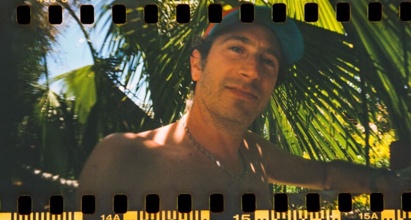 Un hombre sin camisa y con un sombrero azul se encuentra bajo una hoja de palma, mientras la luz del sol se filtra a través de las hojas. La imagen tiene orificios para las ruedas dentadas de la película visibles en la parte superior e inferior, lo que le da un aspecto vintage.