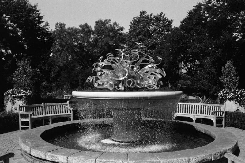 A large fountain with a sculptural glass centerpiece sits surrounded by benches and trees in a landscaped garden. Water cascades from the fountain into a circular stone basin. The image is in black and white.