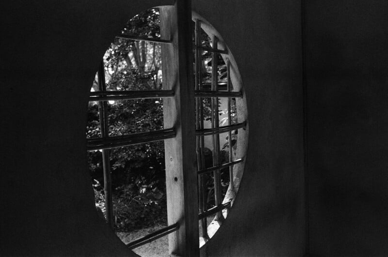 Black and white photo of a round window with wooden grilles, partially open, revealing trees and foliage outside. Shadows and light create a dramatic, tranquil atmosphere inside the space.