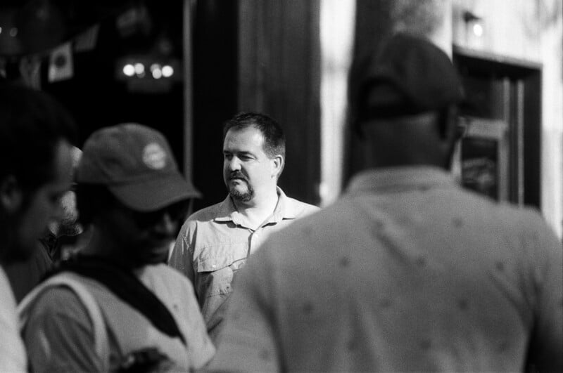 A man with a mustache and goatee stands in the sunlight, surrounded by people in the foreground, some wearing hats. The scene is in black and white with a blurred background.