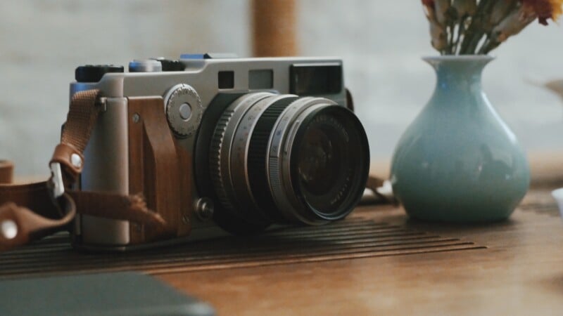 A vintage-style camera with a brown leather strap rests on a wooden surface next to a small blue vase holding dried flowers.