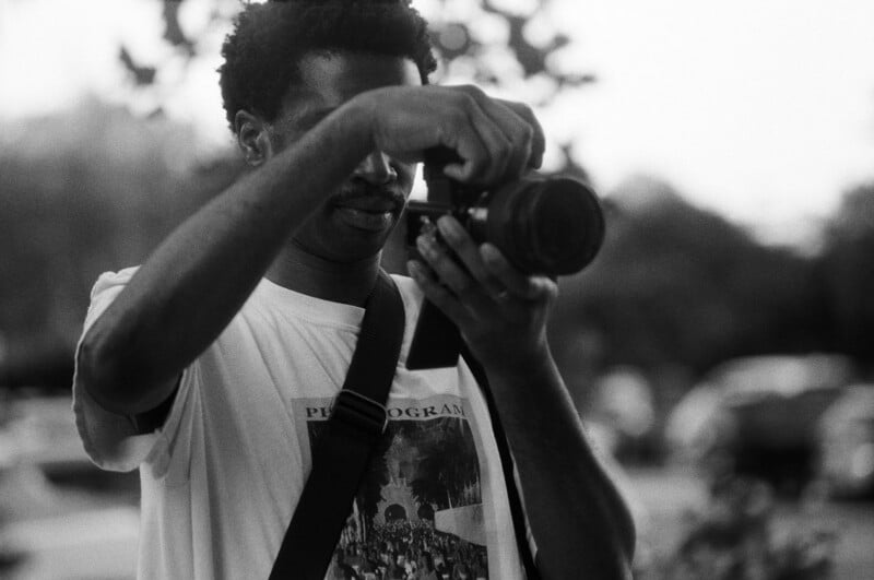 A person wearing a printed T-shirt and shoulder strap adjusts a camera outdoors, with blurred trees and cars in the background. The image is in black and white.