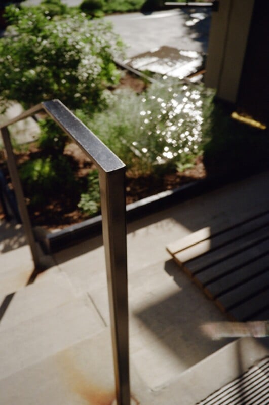 A close-up of a metal handrail on stone steps, with sunlight reflecting off greenery and water in the background, casting shadows on the steps.