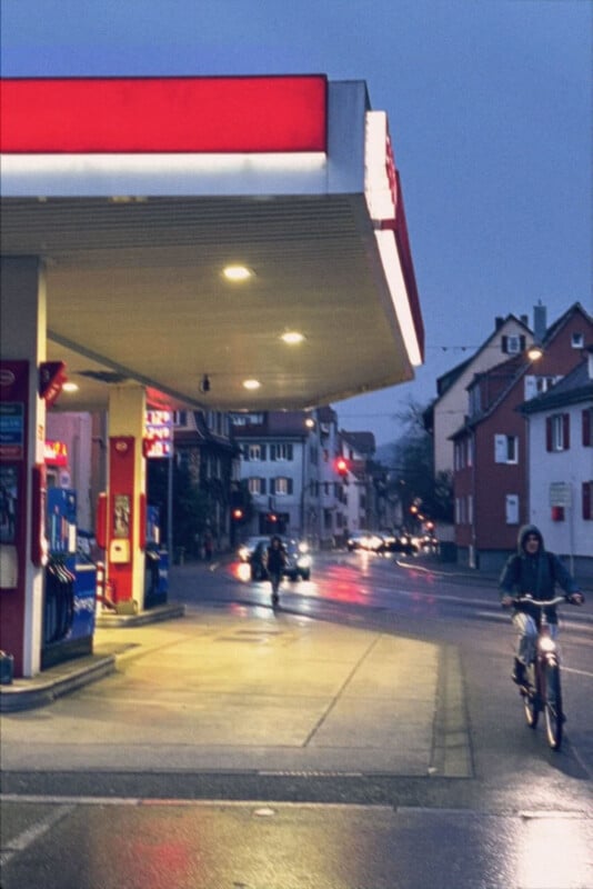 A person rides a bicycle past a gas station on a wet street at dusk, with cars and pedestrians in the background and buildings lining both sides of the road.