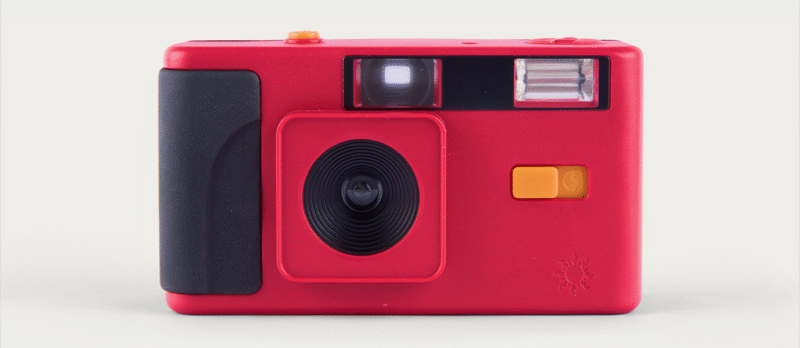 A red, rectangular analog camera with a black grip on the left, a large black lens in the center, a yellow shutter button on top, a yellow switch on the front, and flash components next to the viewfinder.