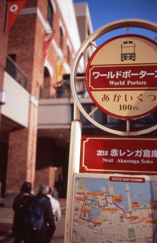 A close-up of a circular tram stop sign for "World Porters" with Japanese and English text, in front of a brick building; a map is visible below and people walk in the background.