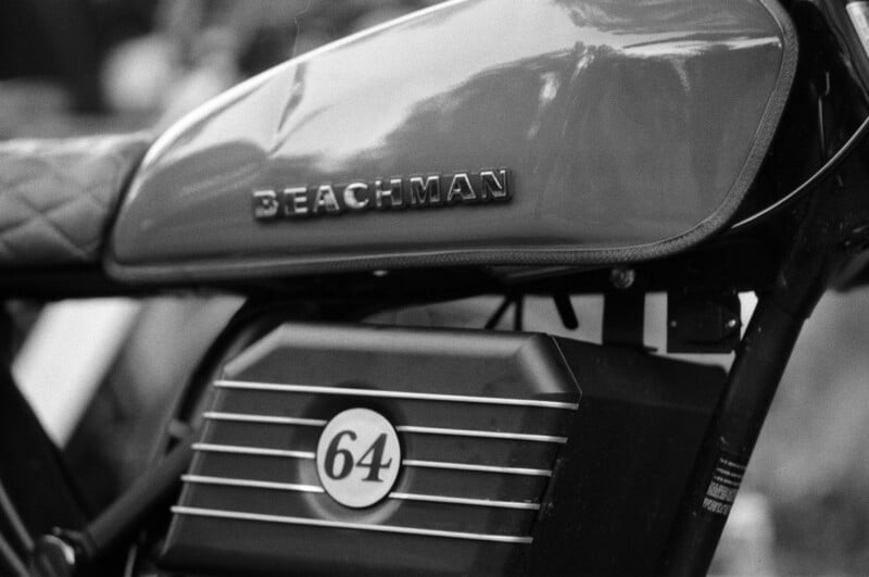 Black and white close-up of a motorcycle tank labeled "BEACHMAN," with the number 64 on a panel below it.