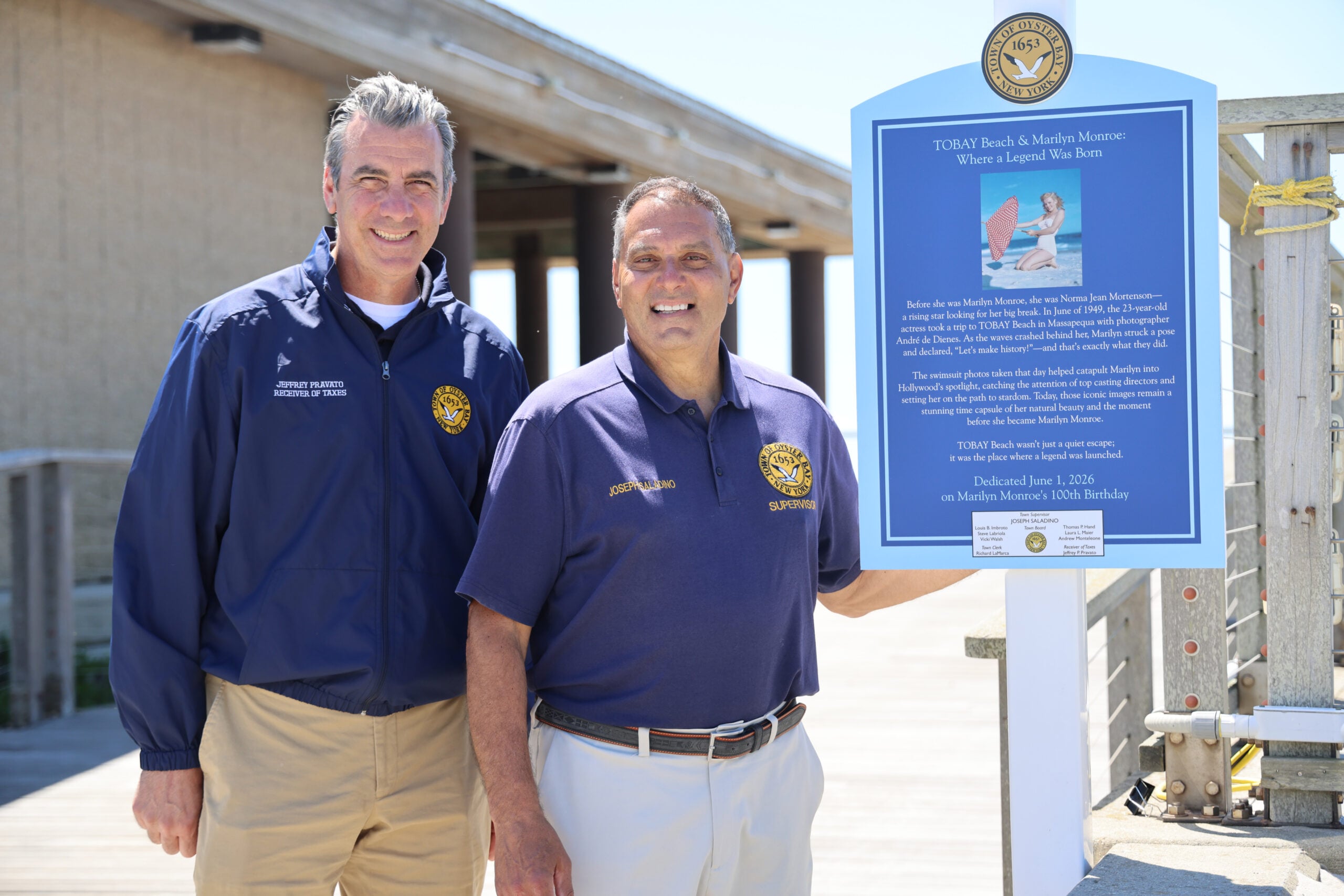 Two men stand smiling next to a blue outdoor informational sign about a marine museum. Both wear official uniforms with badges. The background includes a wooden structure and clear blue sky.