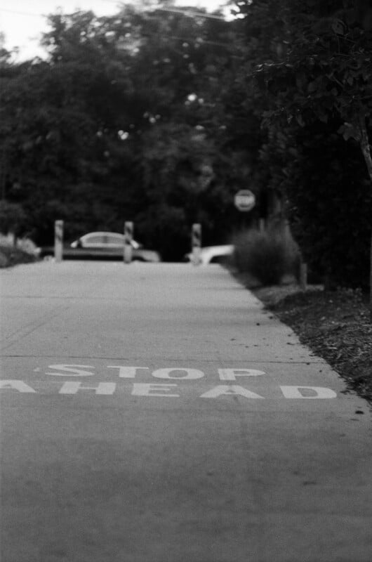 Black-and-white photo of a sidewalk with the words "STOP AHEAD" painted on it. In the background, there are trees, parked cars, and a stop sign partially visible.