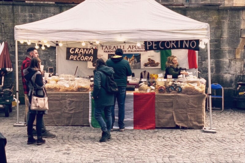 A market stall under a white tent sells Italian cheeses and snacks. Signs read “GRANA DI PECORA” and “PECORINO.” Three people stand in front, and an Italian flag hangs on the table.