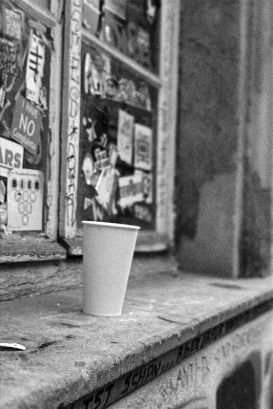 A white paper cup sits on a ledge beneath a window covered in various colorful stickers and posters. The scene is in black and white, giving it a vintage or urban feel.