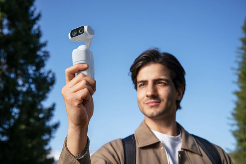 A man outdoors holds up a white handheld camera or gimbal, looking at it with a slight smile. The sky is clear and blue, with tall trees in the background.
