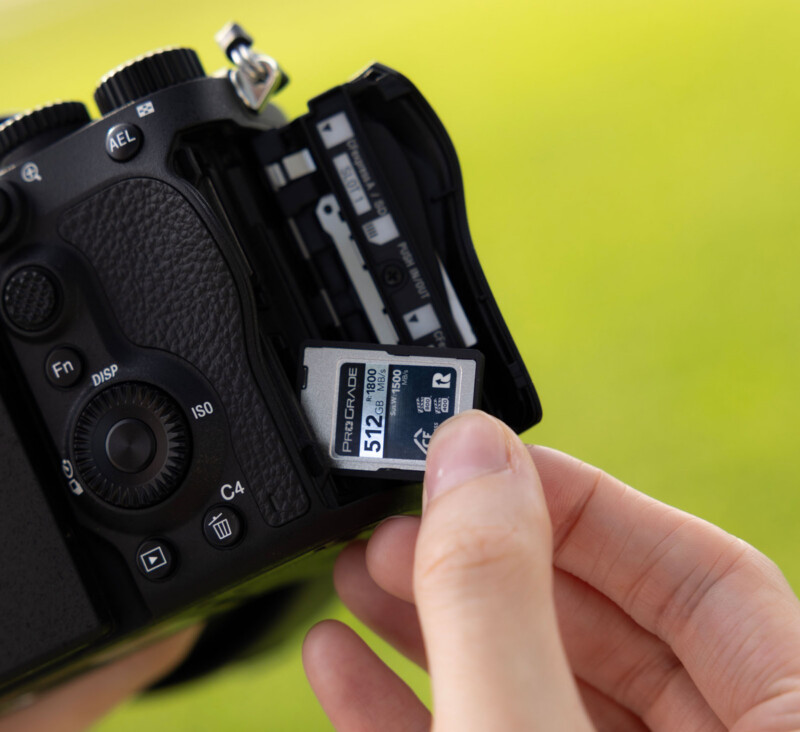 A hand inserts a 512GB ProGrade SD memory card into the open memory card slot of a black digital camera, with a blurred green background.