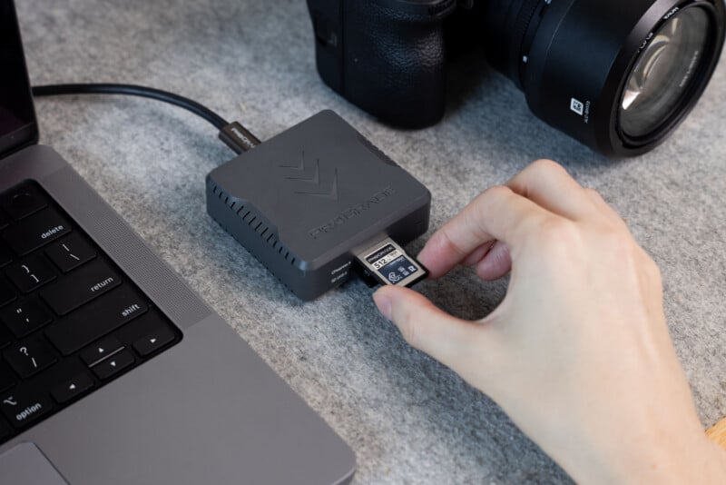 A hand inserts an SD card into a gray card reader connected to a laptop, with a camera and lens visible in the background.