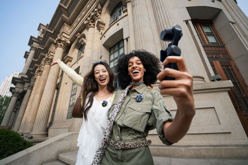 Two young women stand on stone steps in front of a historic building, smiling and posing for a selfie with a handheld camera. One has her arm around the other, and both look happy and excited.