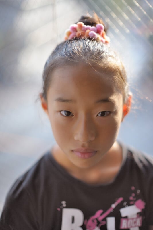 A young girl with her dark hair tied up in a bun decorated with a pink scrunchie looks directly at the camera, standing in soft natural light. She wears a black t-shirt with white and pink lettering.