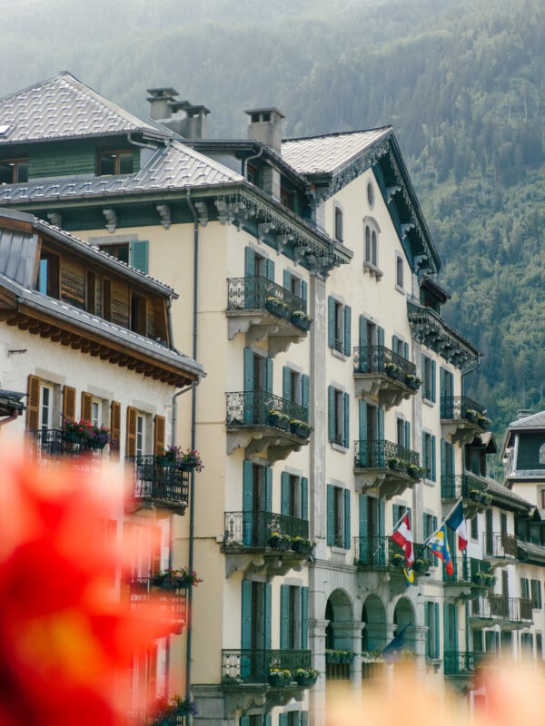 A picturesque European building with green shutters and balconies, flying several flags, set against a backdrop of misty, forested mountains and blurred red flowers in the foreground.
