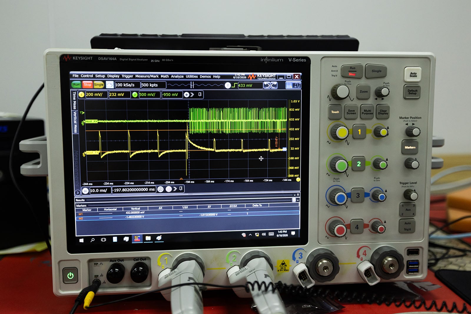 A digital oscilloscope displays a waveform with green and yellow traces on its screen. The device features multiple buttons, dials, and input ports below the display, with cables connected to the front panel.