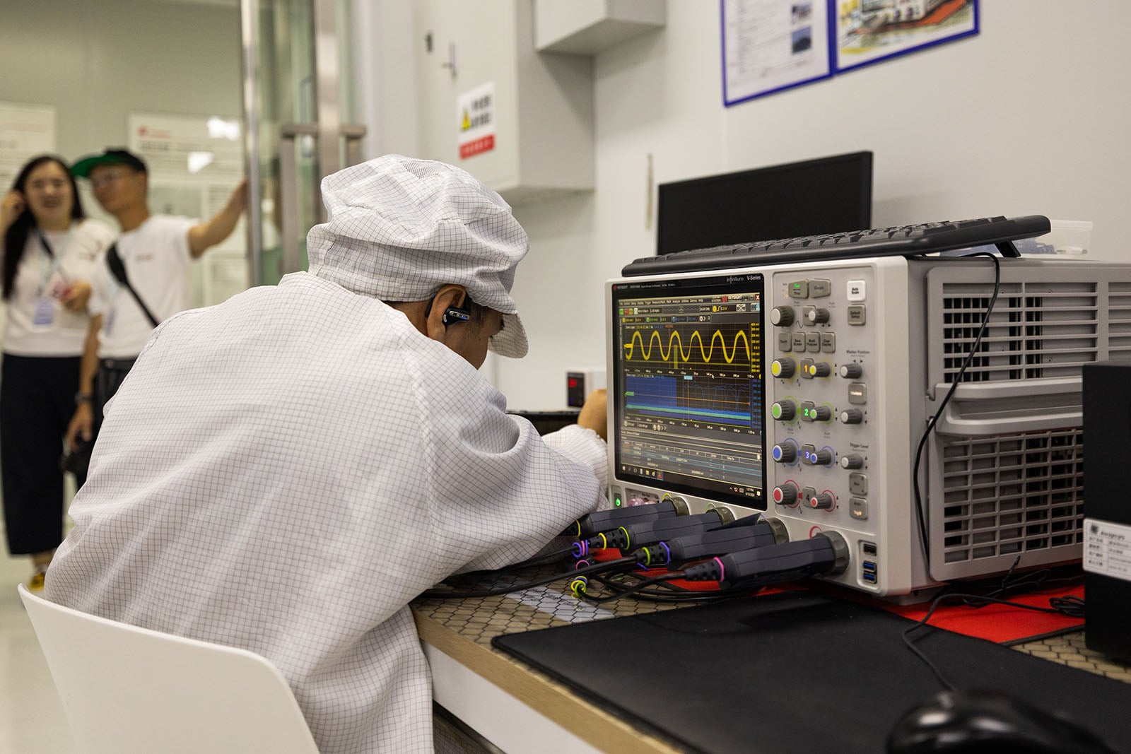 A person in a lab coat and cap operates an oscilloscope displaying waveforms, while three people observe from the background in a clean laboratory setting.